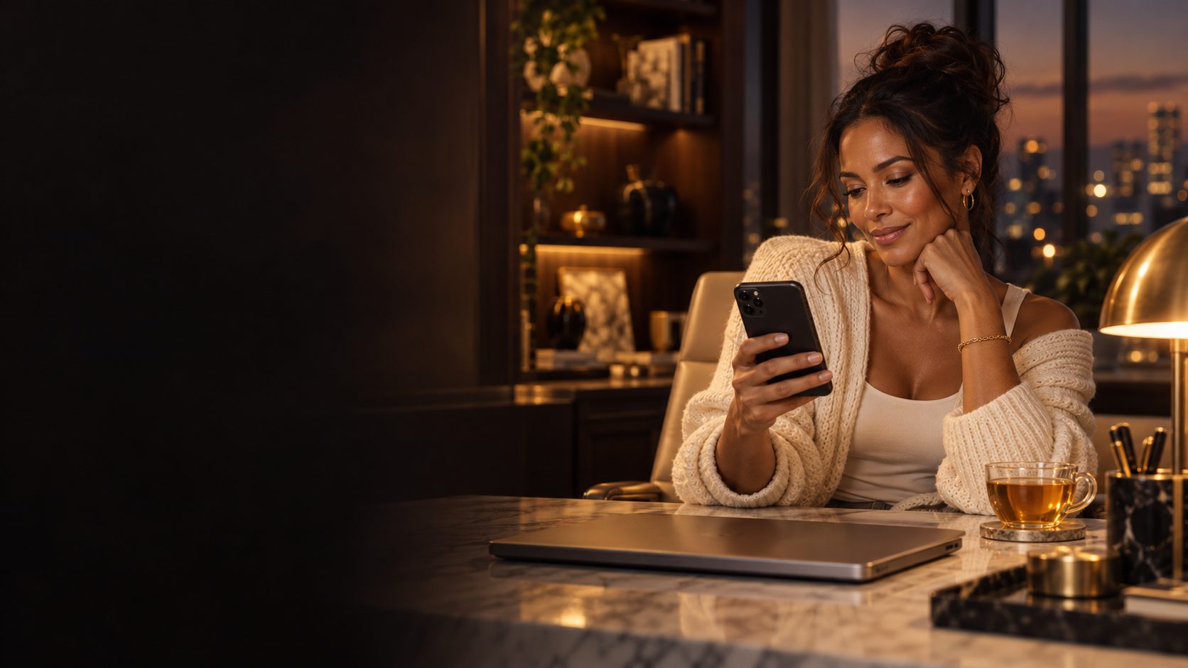 A woman in a cream cardigan smiles at her phone at a marble desk in the evening, city lights behind her